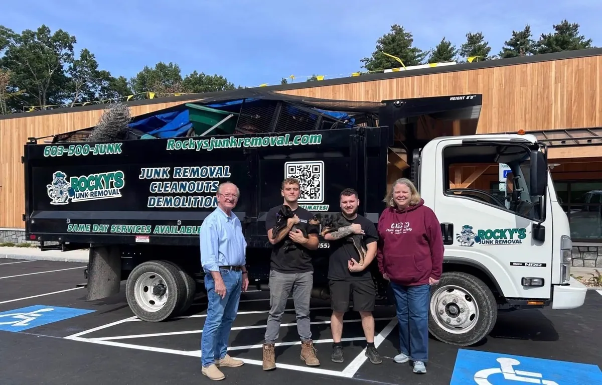 Rocky's Junk Removal crew holding two puppies with Humane Society for Greater Nashua staff in front of a Rocky's truck loaded with fencing, tarps, and scrap from the shelter